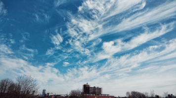 A blue, cloud-swept Manchester sky with trees and buildings at the bottom of the frame