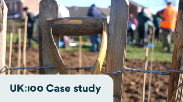 A spade lent against a wood and wire fence with a community of people out of focus in the background having just planted trees.